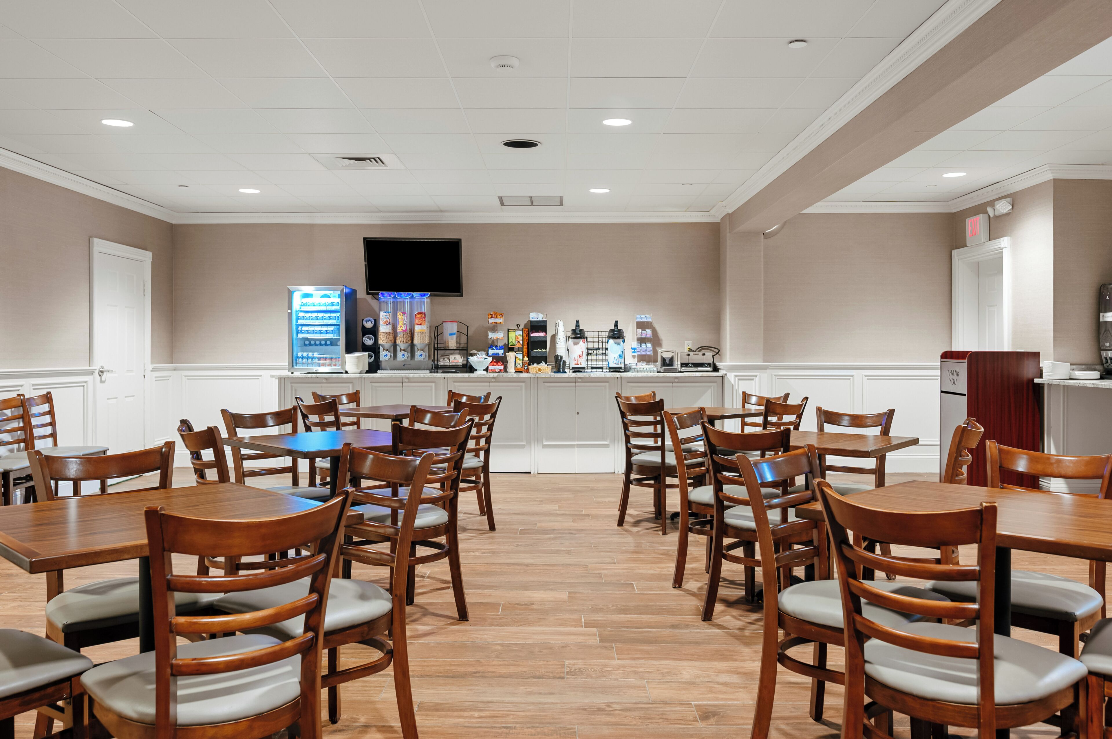 A hotel dining area with multiple wooden tables and chairs, a beverage station, and a flat-screen TV mounted on the wall.