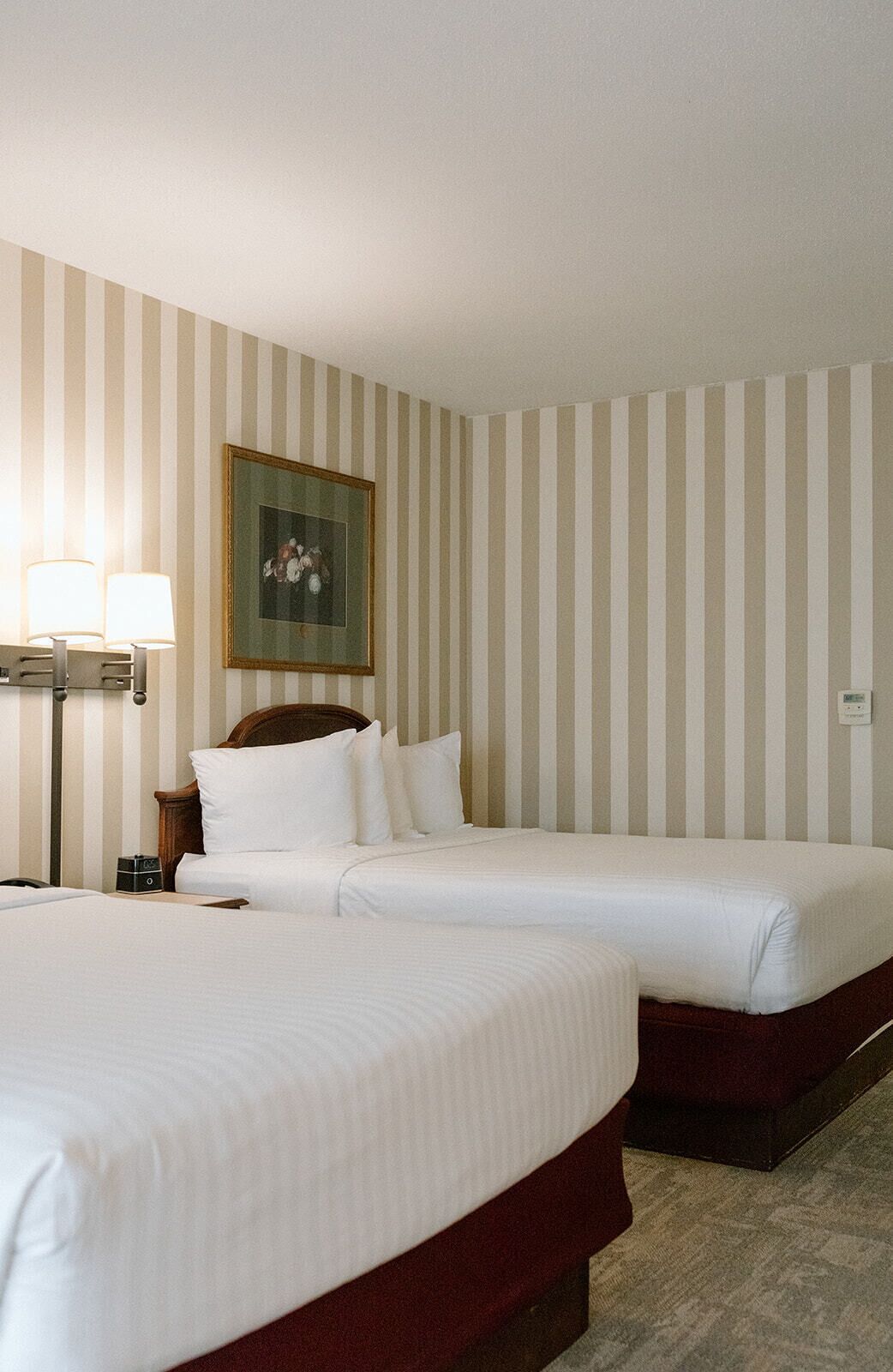 A hotel room with two beds, a nightstand, a lamp, and a painting on the wall.
