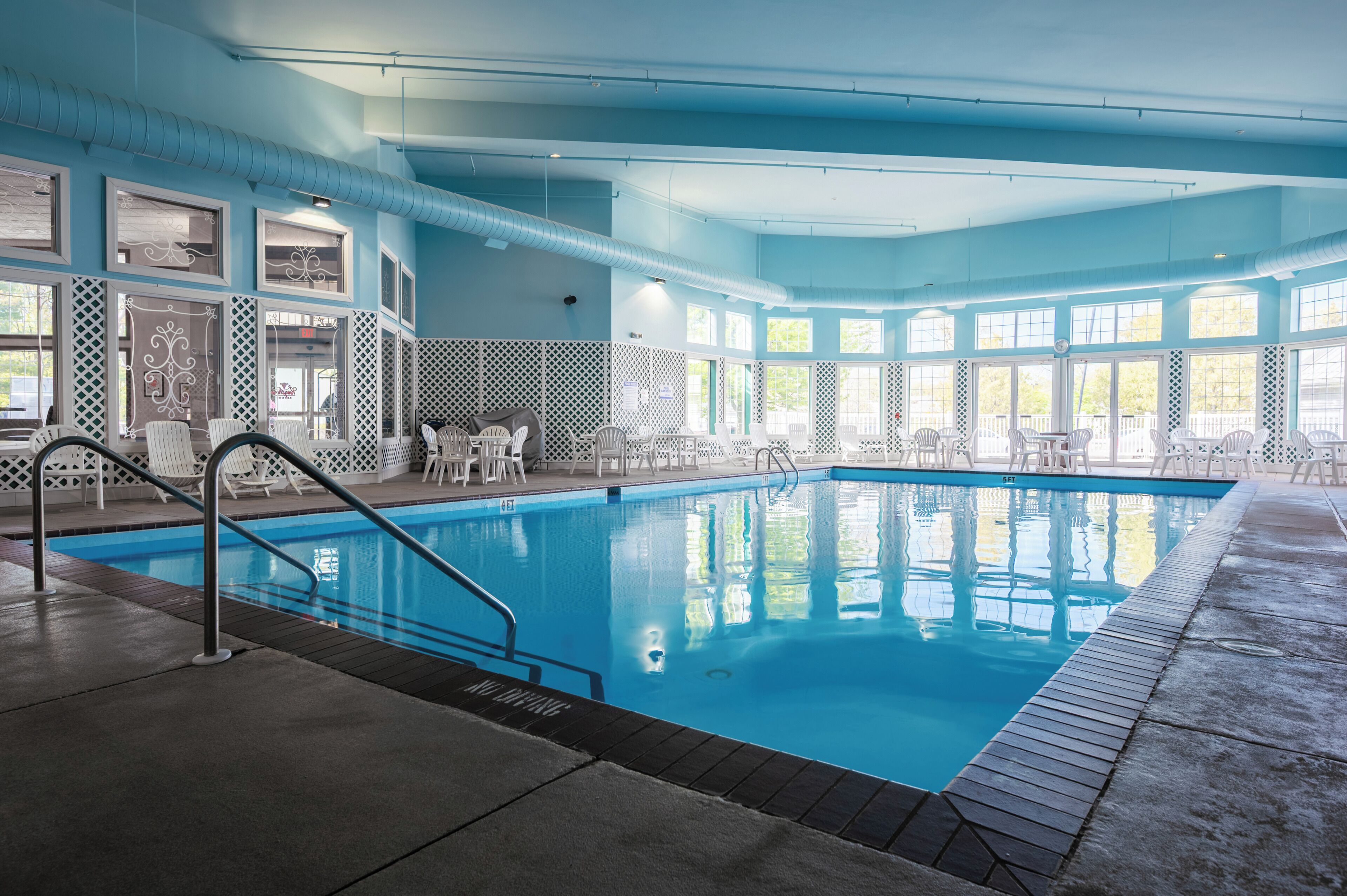 An indoor swimming pool with clear blue water, surrounded by white chairs and tables, and large windows allowing natural light.