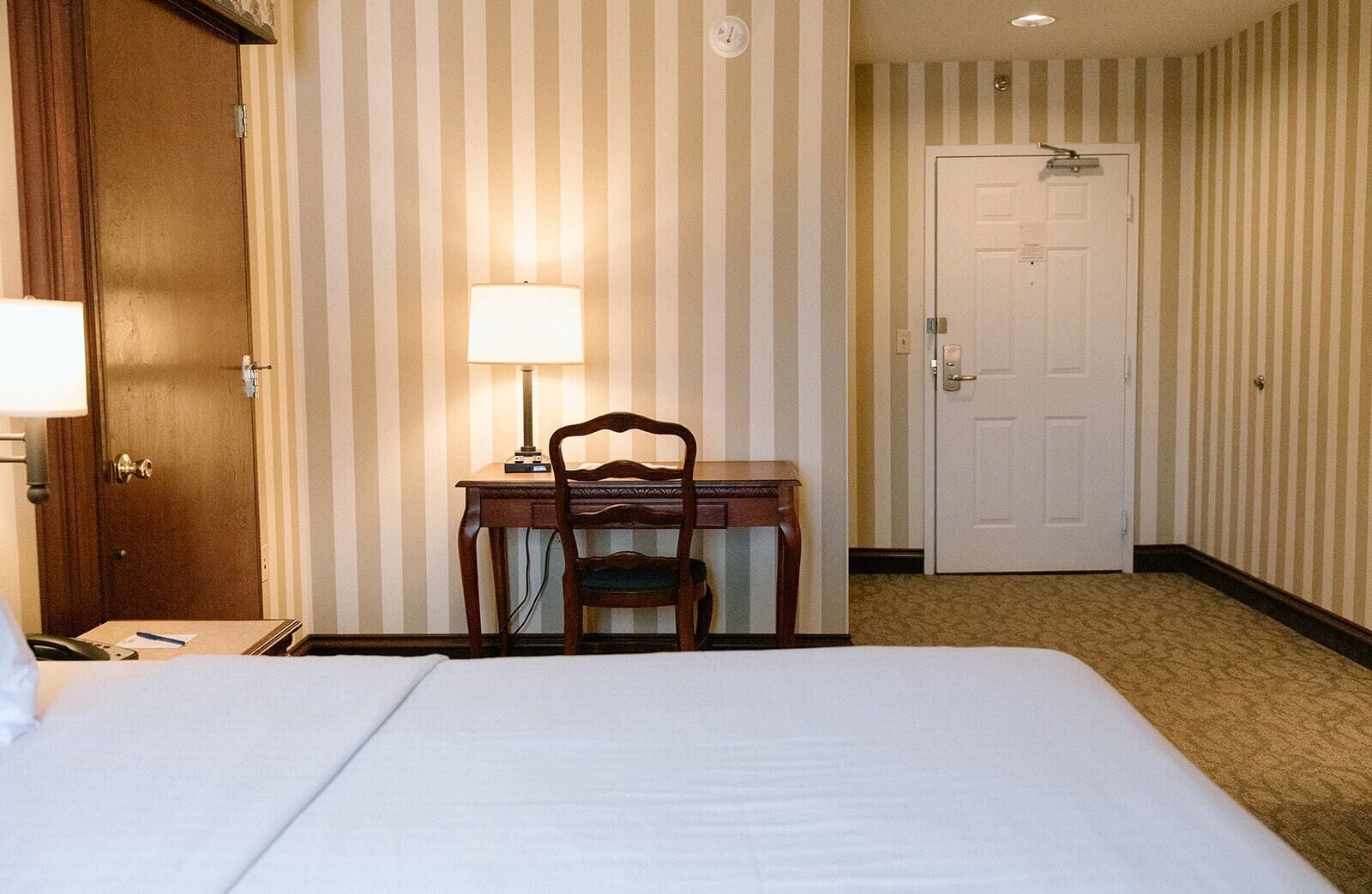 Une chambre d’hôtel avec un lit, un bureau et une chaise, une lampe, une porte et un papier peint à rayures.