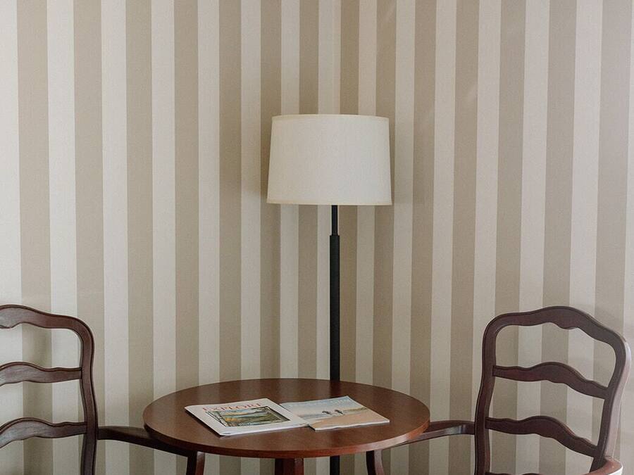 A corner room with striped wallpaper, a round wooden table with two chairs, a floor lamp, and a magazine on the table.