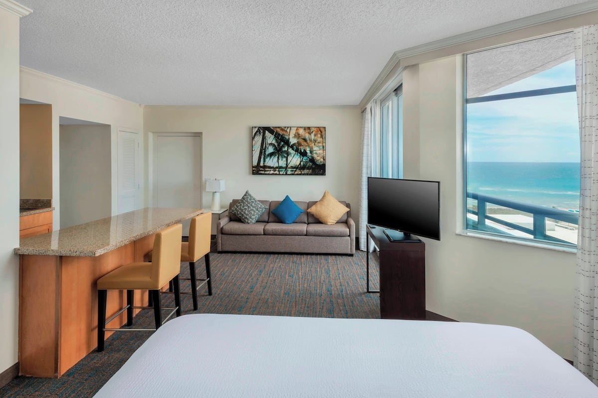 A hotel room with a bed, a sofa, a TV, a desk with chairs, and a view of the ocean.