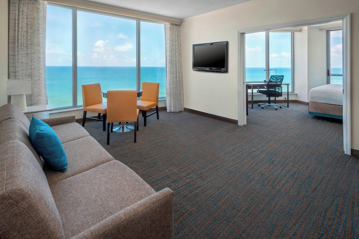 A hotel room with a large window, a flat-screen TV, a sofa, a dining table with chairs, and a bed.