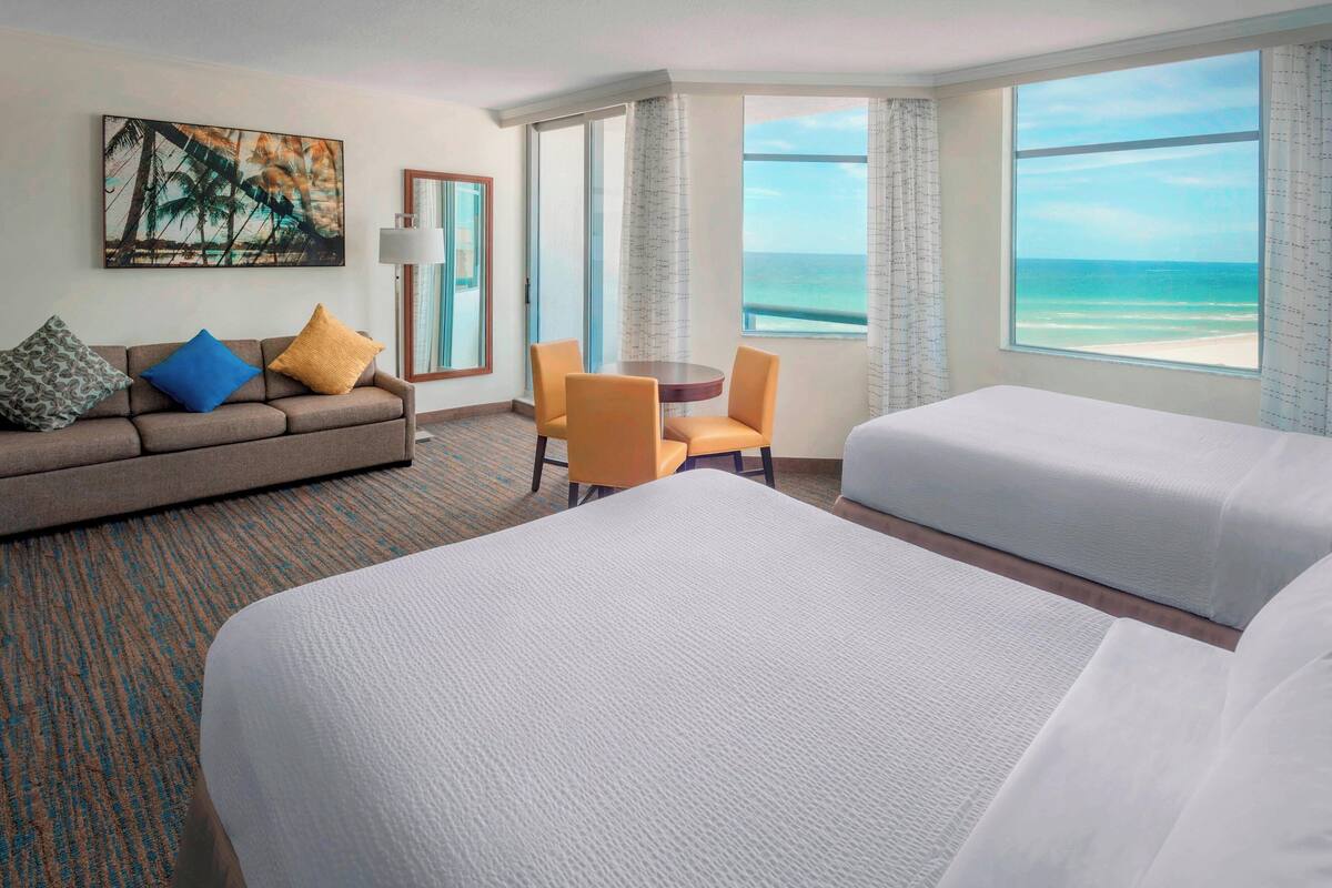 A hotel room with a large bed, a sofa with cushions, a small table, and a view of the ocean.