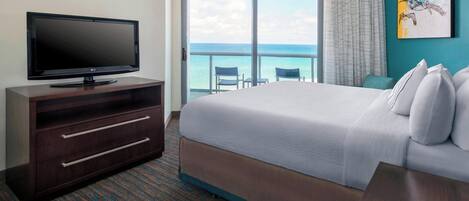 A hotel room with a large bed, a flat-screen TV on a wooden dresser, a balcony with a view of the ocean, and a painting on the wall.