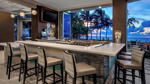 A bar area with a long counter, multiple bar stools, and a view of the ocean through large windows. - Residence Inn Fort Lauderdale Pompano Beach / Oceanfront (Pompano Beach)