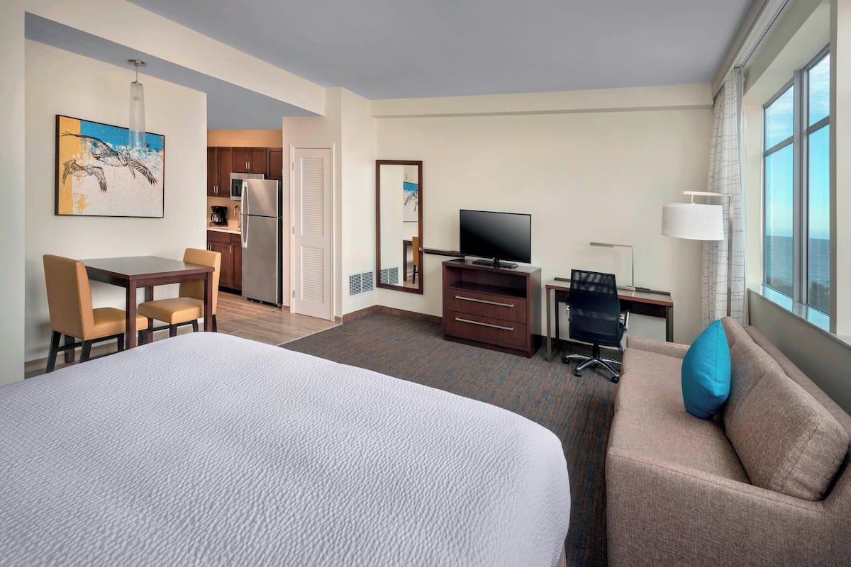 A hotel room with a large bed, a sofa with a blue pillow, a desk with a computer, a television, and a dining area with chairs.