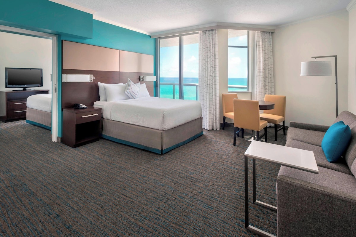 A hotel room with a large bed, a desk, a chair, a TV, and a view of the ocean.