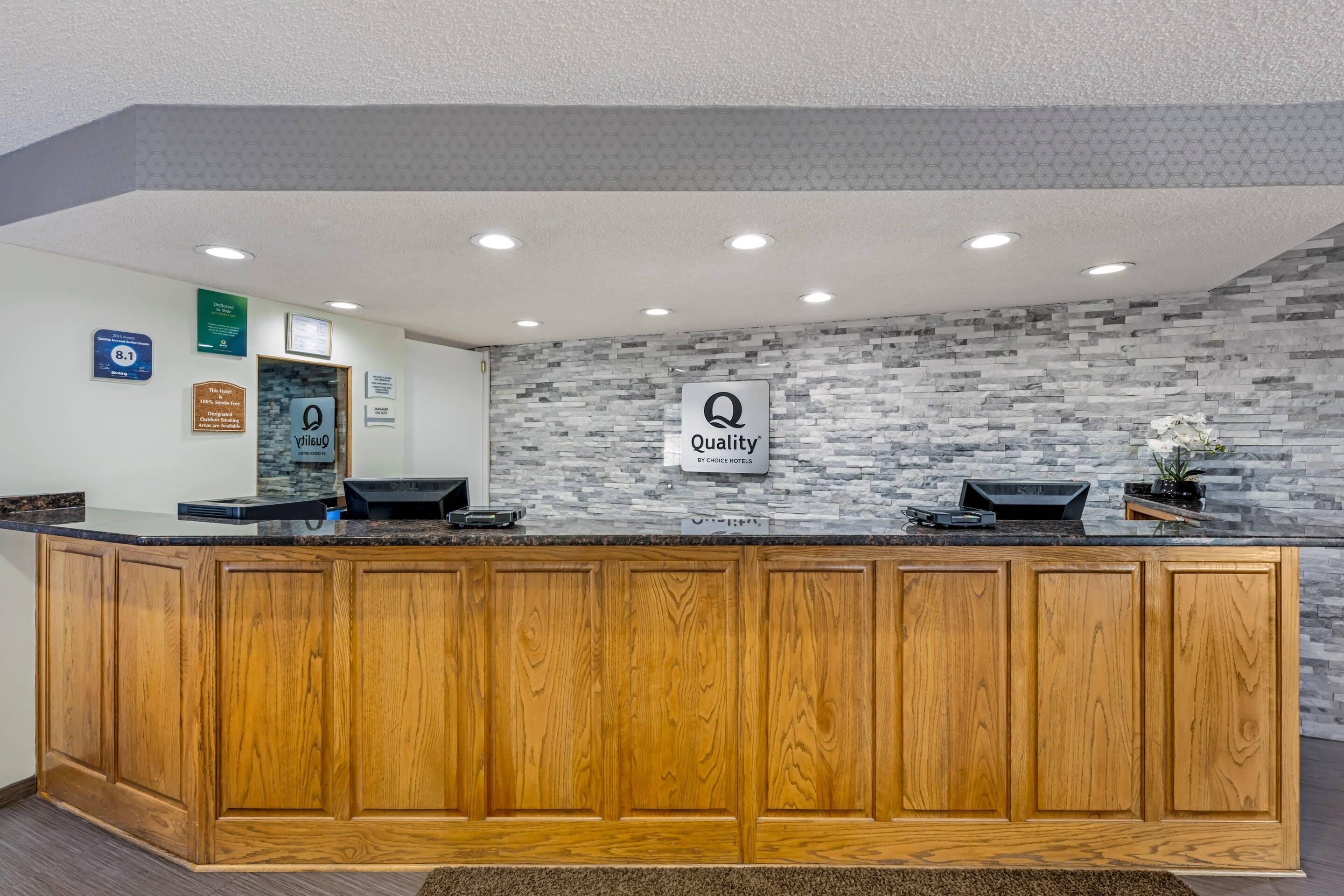 Un comptoir d’accueil avec un mur en pierre en arrière-plan, une enseigne indiquant « Qualité, une promesse de Choice Hotels », et une plaque commémorative du « Prix de la qualité 2015 ».