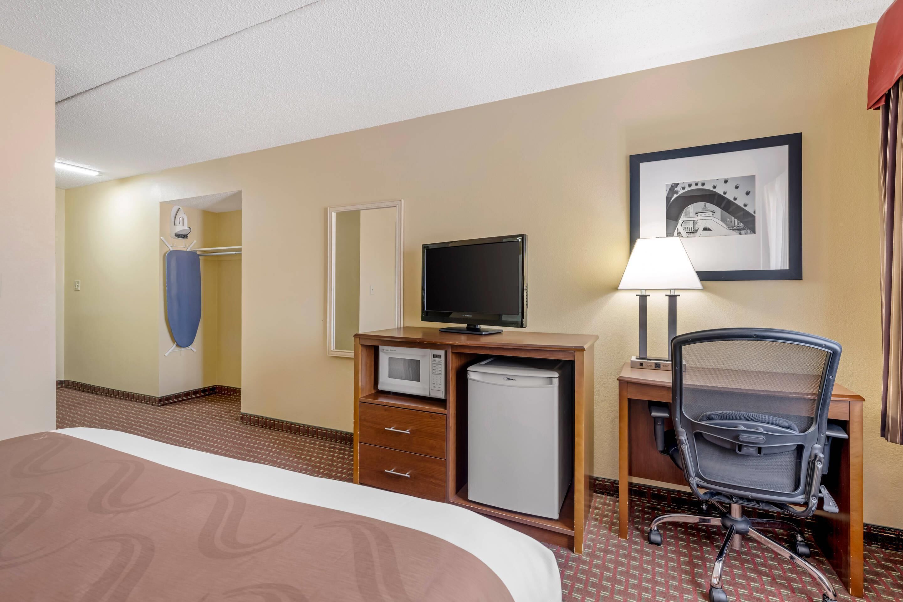 A hotel room with a bed, a desk with a computer, a chair, a microwave, and a television.