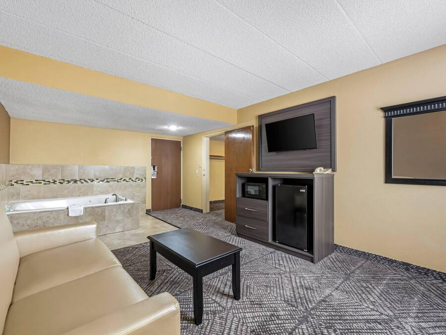 A hotel room with a sofa, a small table, a flat-screen TV, a minibar, and a bathroom with a bathtub.