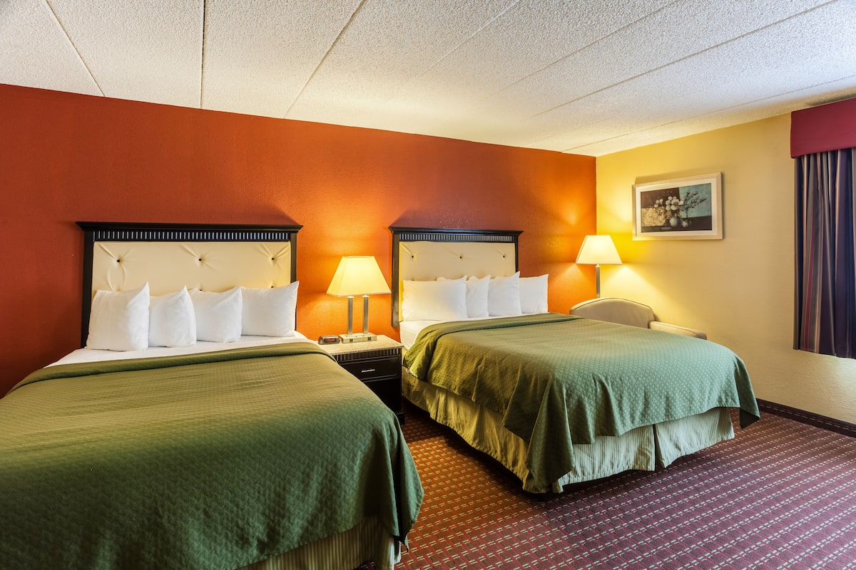 Une chambre d’hôtel avec deux lits, chacun recouvert d’une couvre-lit verte et agrémenté d’oreillers blancs. Une table de chevet avec une lampe se trouve entre les deux lits. Les murs de la chambre sont de couleur orange et le sol est recouvert d’un tapis à motifs.