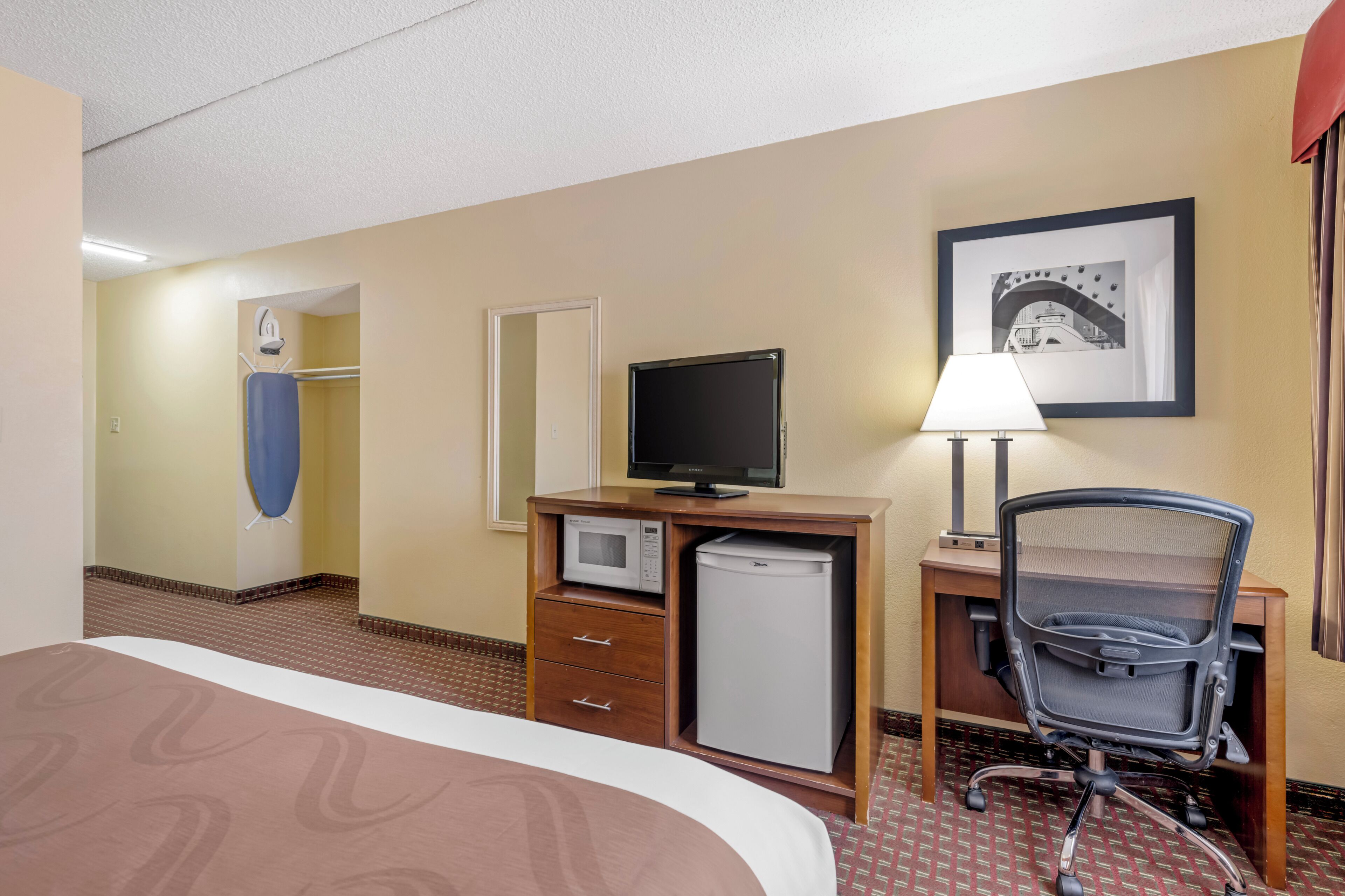 Quarto de hotel com cama, escrivaninha com computador, micro-ondas, cadeira e quadro na parede.