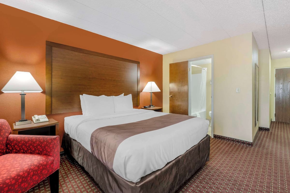 Une chambre d’hôtel avec un grand lit, deux lampes de chevet, un fauteuil rouge et une tête de lit en bois.