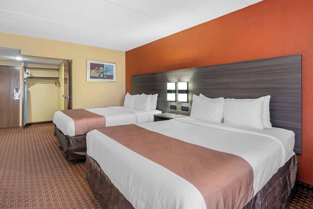 Une chambre d’hôtel avec deux lits, une tête de lit en bois, une moquette marron et une porte dotée d’un interrupteur.