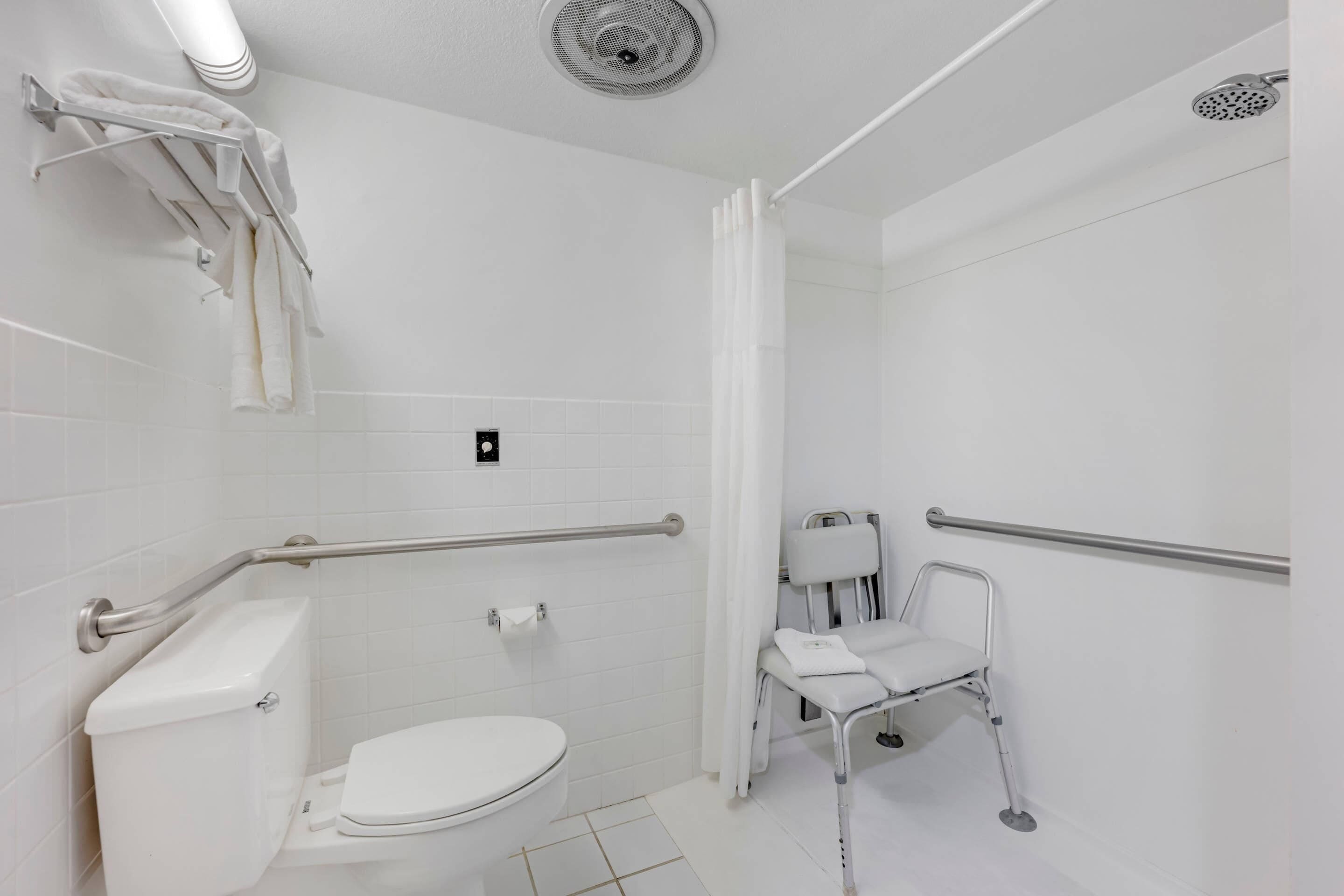 Baño blanco con inodoro, barras de apoyo, zona de ducha y banca accesible para sillas de ruedas.