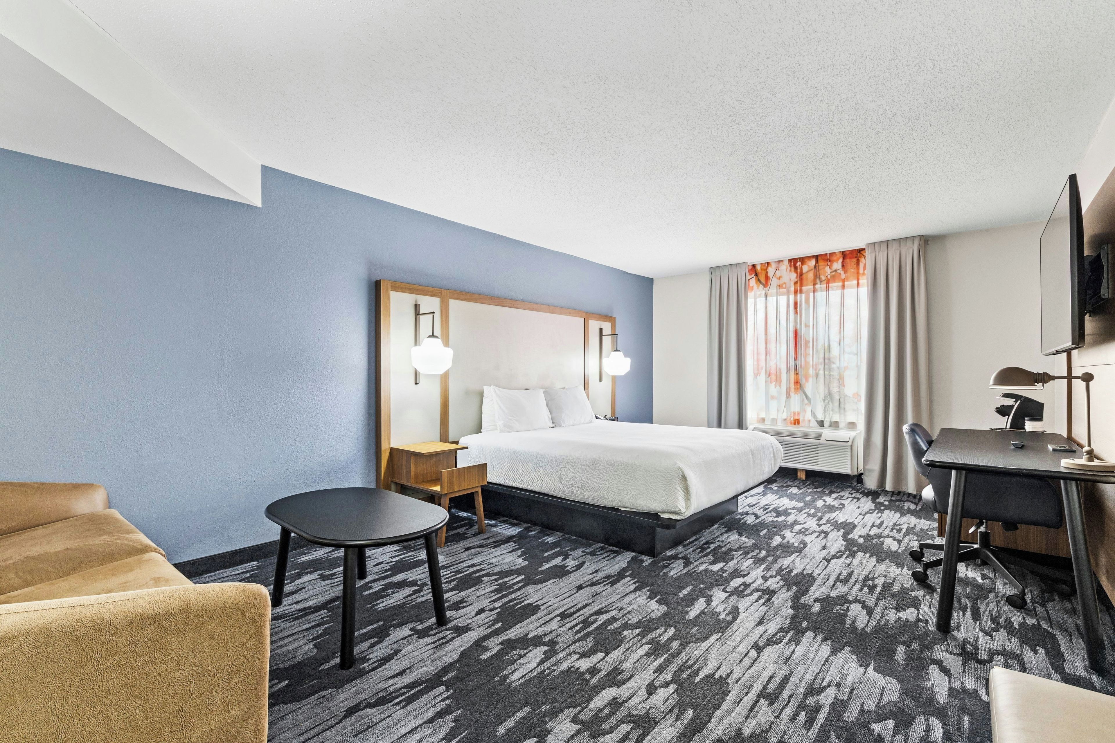 A hotel room with a bed, a desk, a chair, a small table, and a TV.