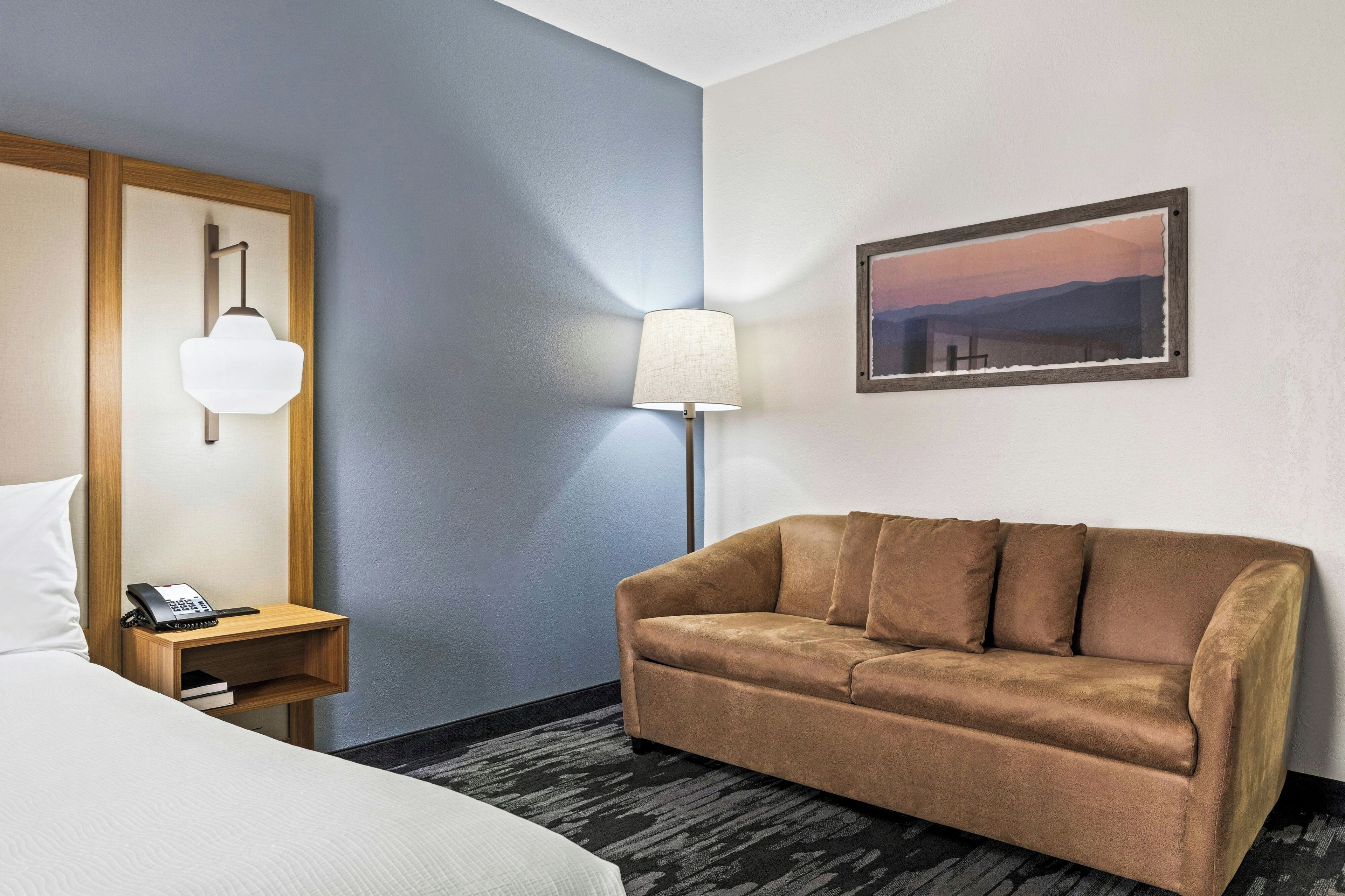A hotel room with a bed, a wooden nightstand with a phone, a brown sofa, a lamp, and a framed picture on the wall.
