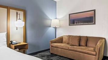 A hotel room with a bed, a wooden nightstand with a phone, a brown sofa, a lamp, and a framed picture on the wall.