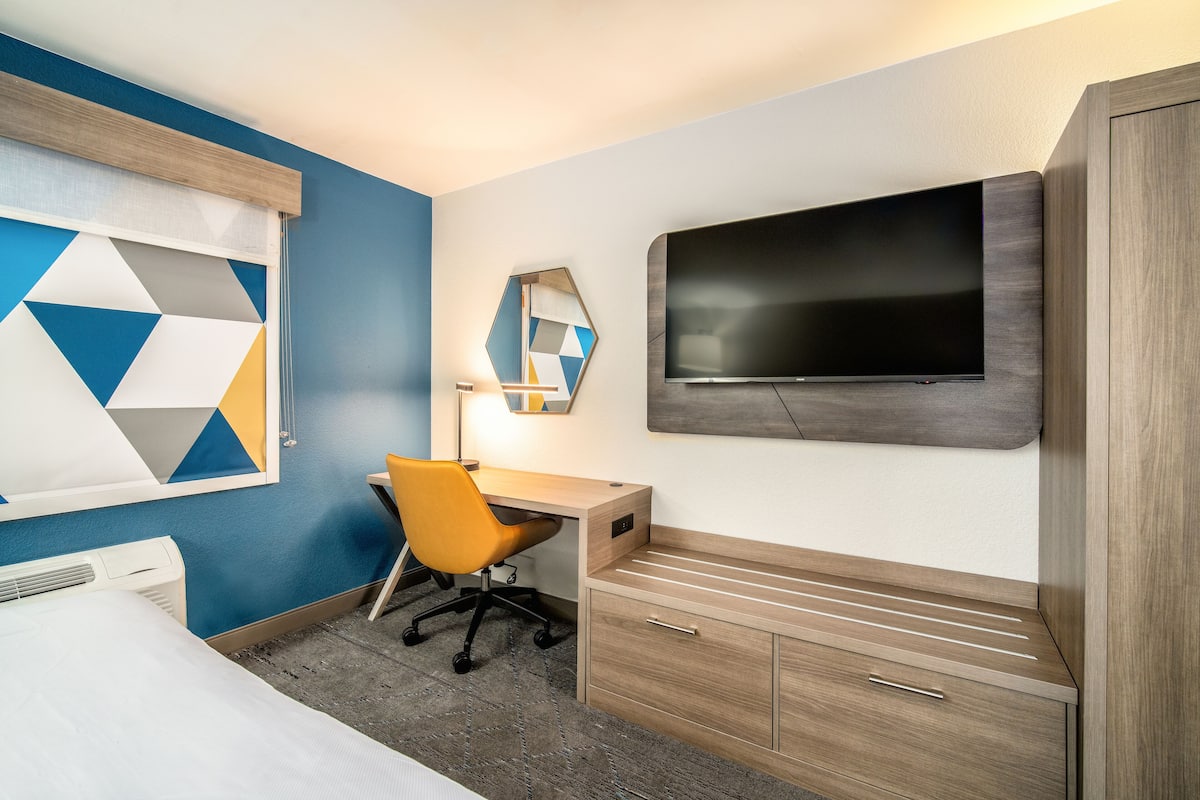 Una habitación de hotel con cama, escritorio con silla, televisor montado en la pared y armario.