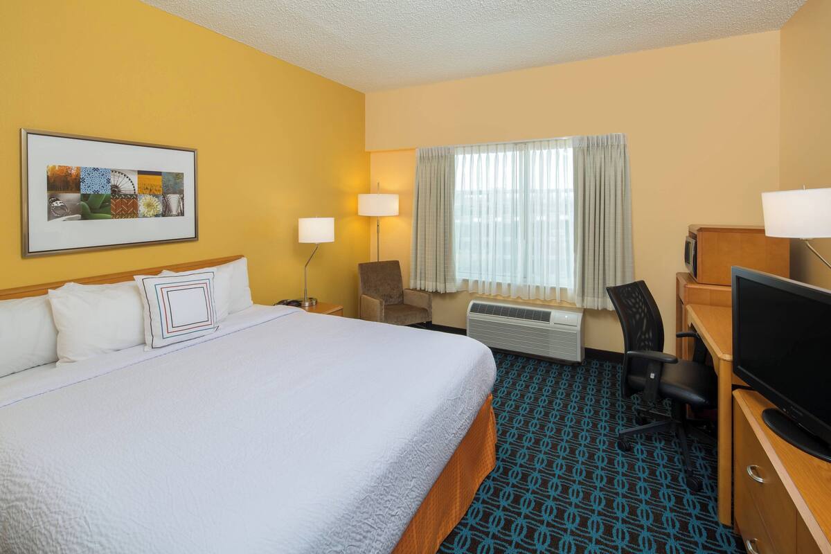 A hotel room with a large bed, a desk with a television, a chair, a desk lamp, a window with curtains, and a framed picture on the wall.