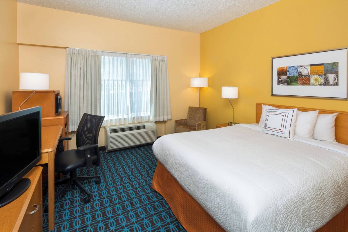 A hotel room with a bed, a desk with a TV, a chair, a desk lamp, a wall-mounted lamp, and a framed picture on the wall.