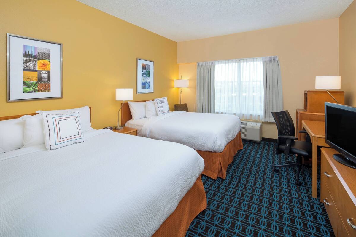 A hotel room with two beds, a desk, a TV, and a window with curtains.