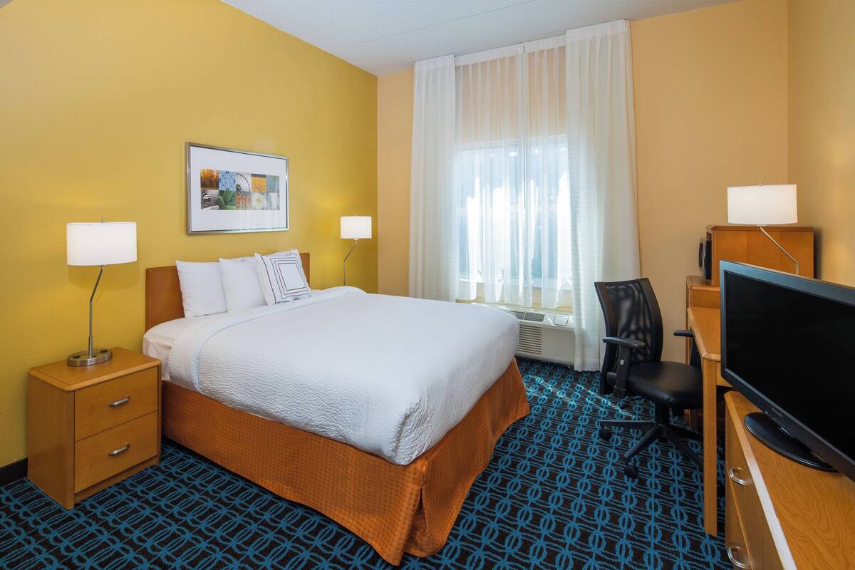 A hotel room with a bed, two bedside tables with lamps, a desk with a computer, a chair, and a large window with curtains.