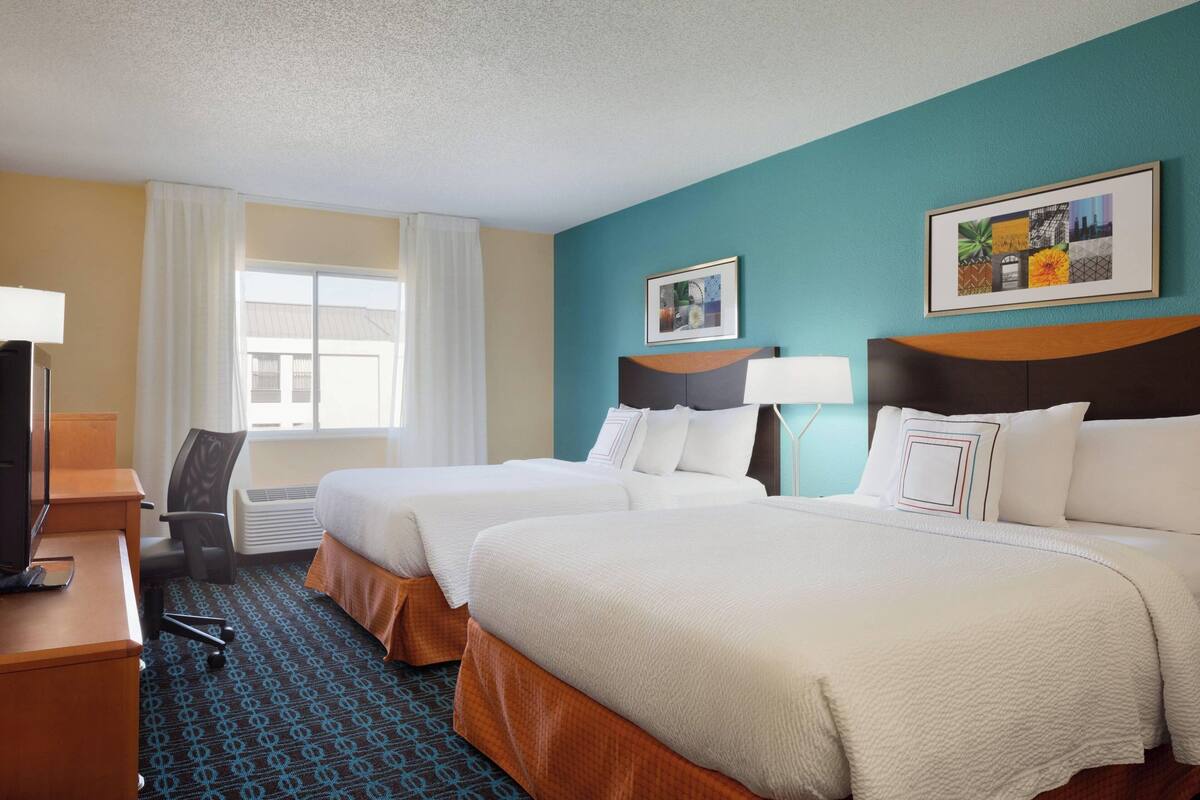 Een hotelkamer met twee bedden, een bureau met een computer, een stoel en een raam met gordijnen.