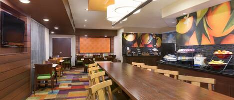 Un restaurante con una larga mesa de madera, sillas, una alfombra colorida y una exhibición de frutas.