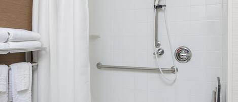 Un área de ducha con pared de azulejos blancos, un cabezal de ducha, una barra de apoyo y un perchero con toallas dobladas.