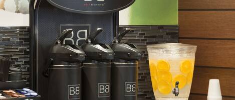 Una estación de bebidas con tres dispensadores etiquetados 'BB' y una jarra grande con rodajas de naranja.