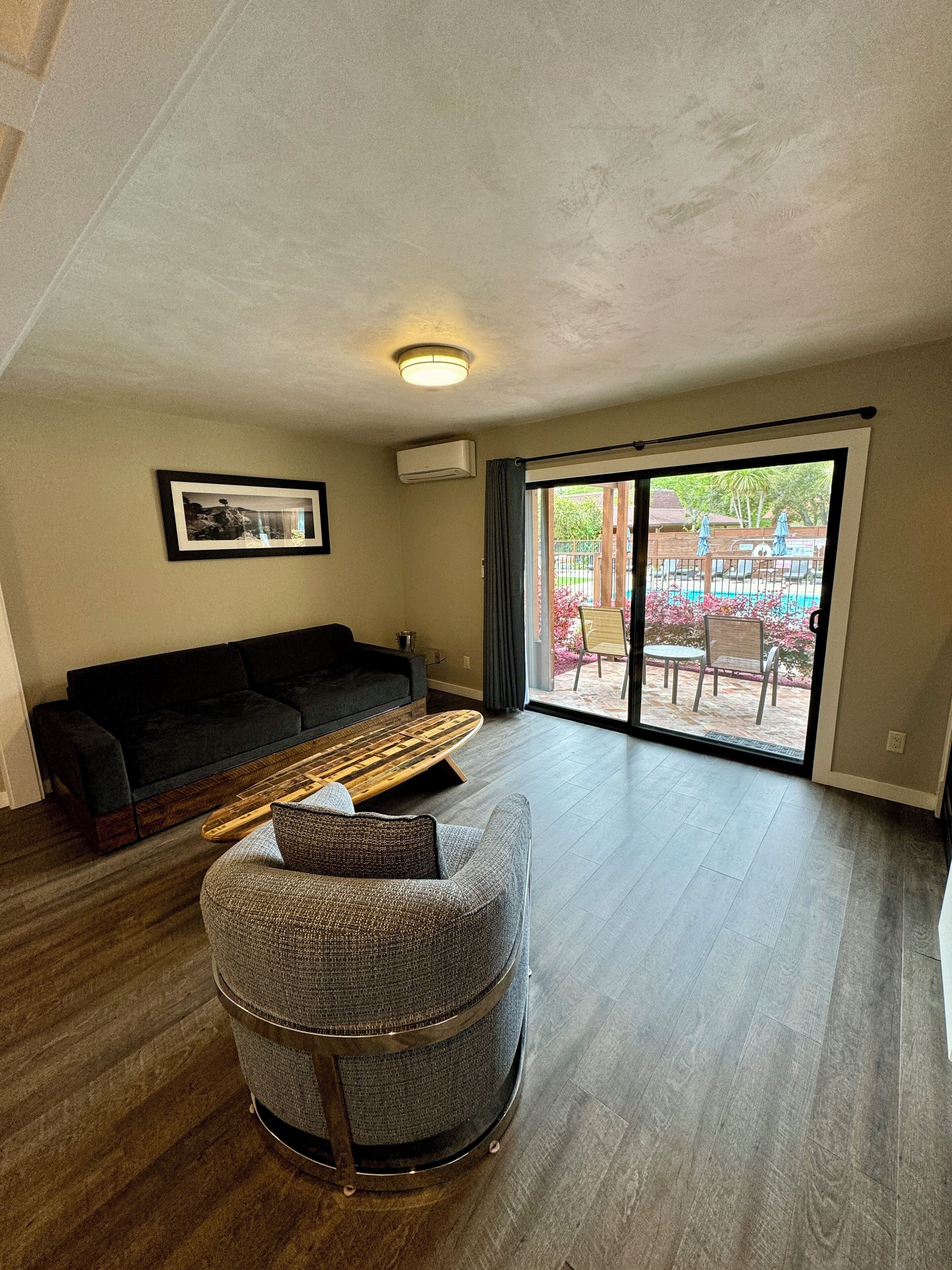A spacious living room with a dark sofa, a round chair, and a wooden coffee table. A large sliding glass door leads to an outdoor area with a pool and patio furniture.