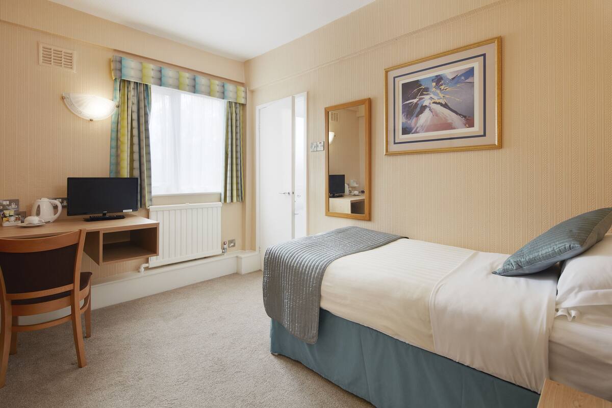 A hotel room with a bed, a desk with a computer, a chair, a window with curtains, a radiator, and a framed picture on the wall.