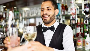 Un barman, vêtu d’un gilet noir et d’un nœud papillon, tient un verre de vin blanc.