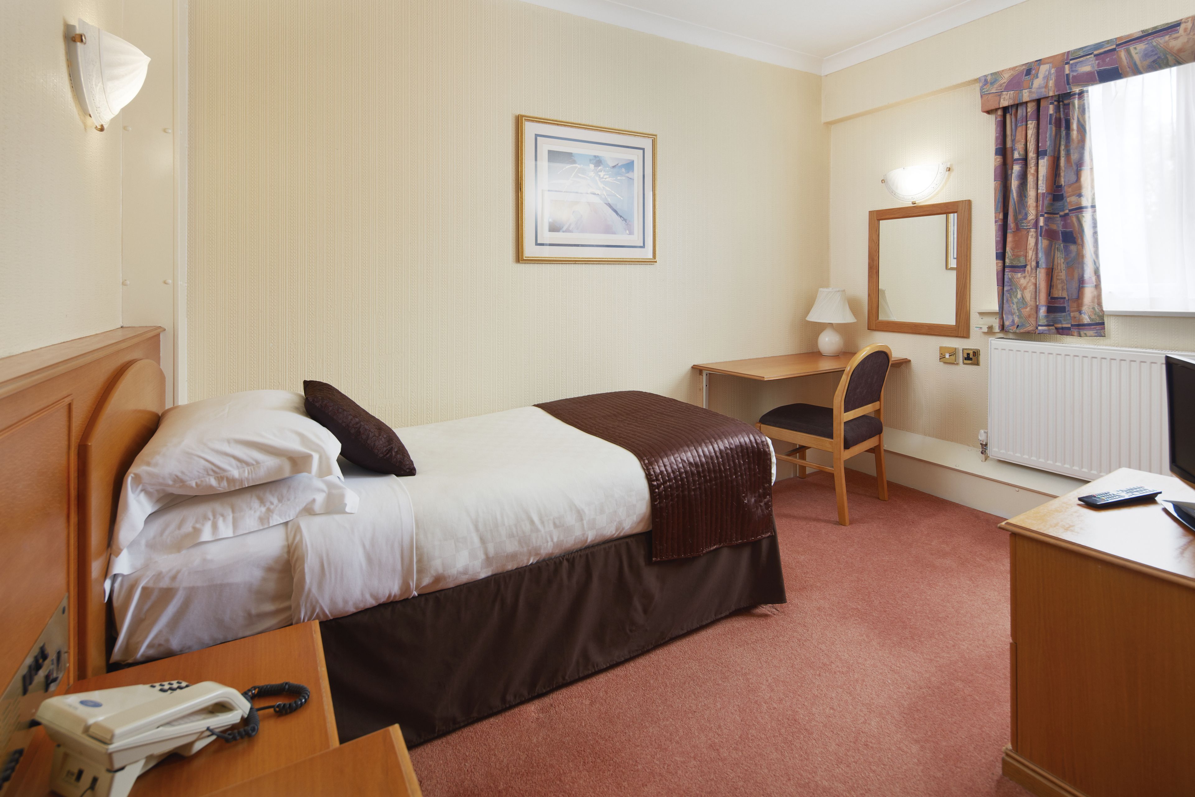 A hotel room with a bed, a desk, a chair, a telephone, a lamp, and a window with curtains.
