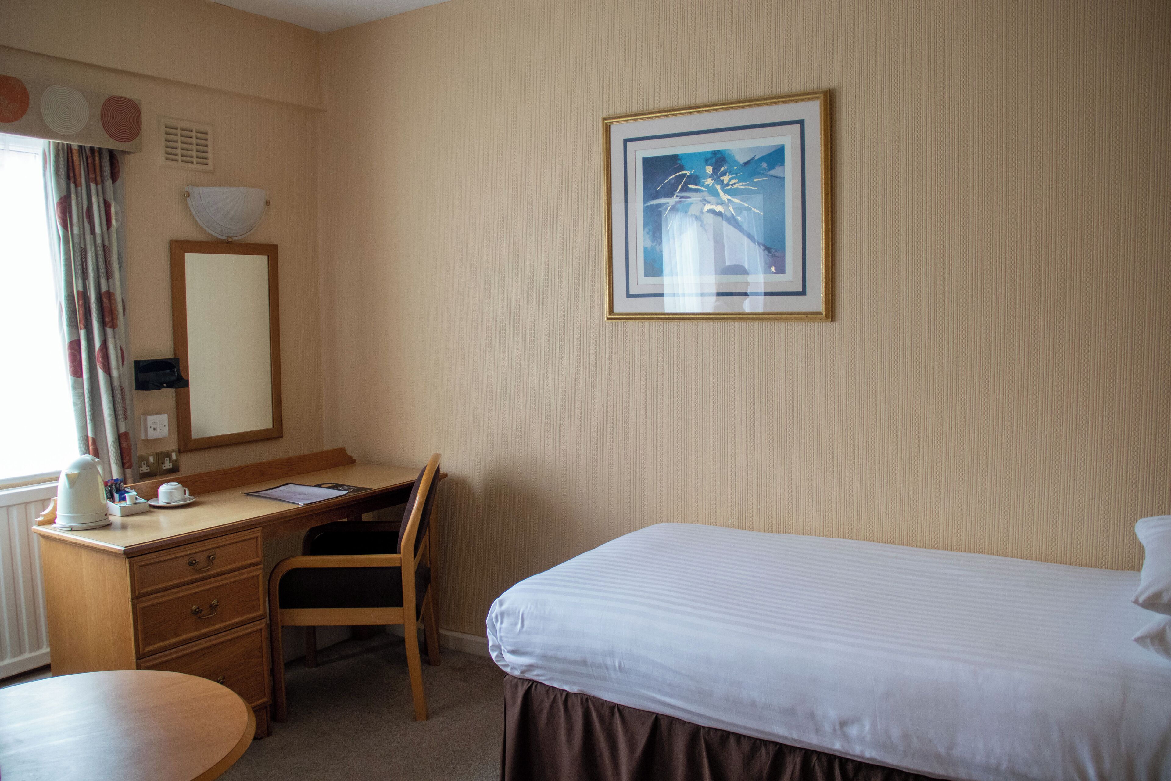 Ein Hotelzimmer mit Bett, Schreibtisch, Stuhl, einem kleinen Tisch, einem Spiegel und einem gerahmten Bild an der Wand.