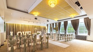 Un lieu de réception pour mariages avec des rangées de chaises, une scène ornée d’une décoration représentant le mot « AMOUR », des lustres et de grandes fenêtres.