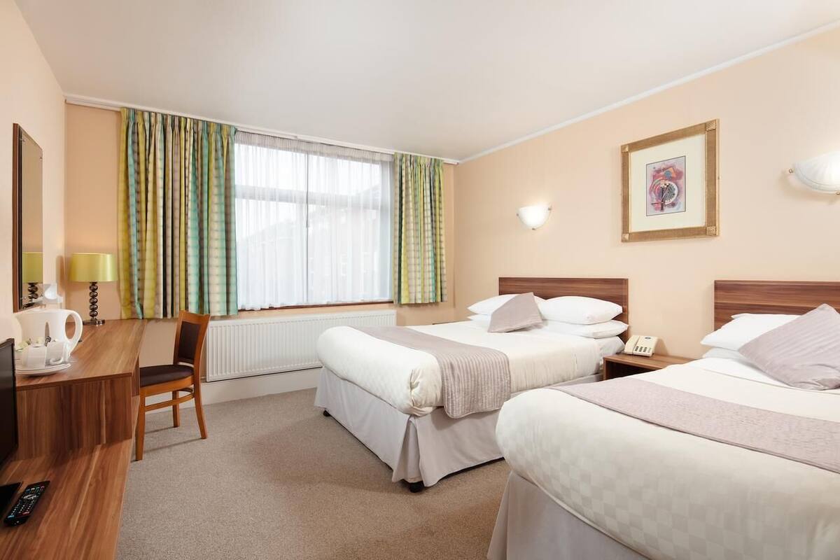 A hotel room with two beds, a desk, a chair, a radiator, a window with curtains, and a wall-mounted light fixture.