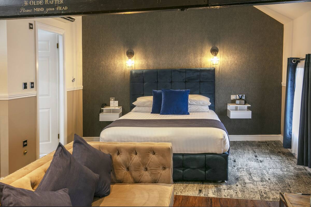 Una habitación de hotel moderna con una cama grande, dos mesitas de noche, un cabecero y una zona de estar con un banco acolchado.