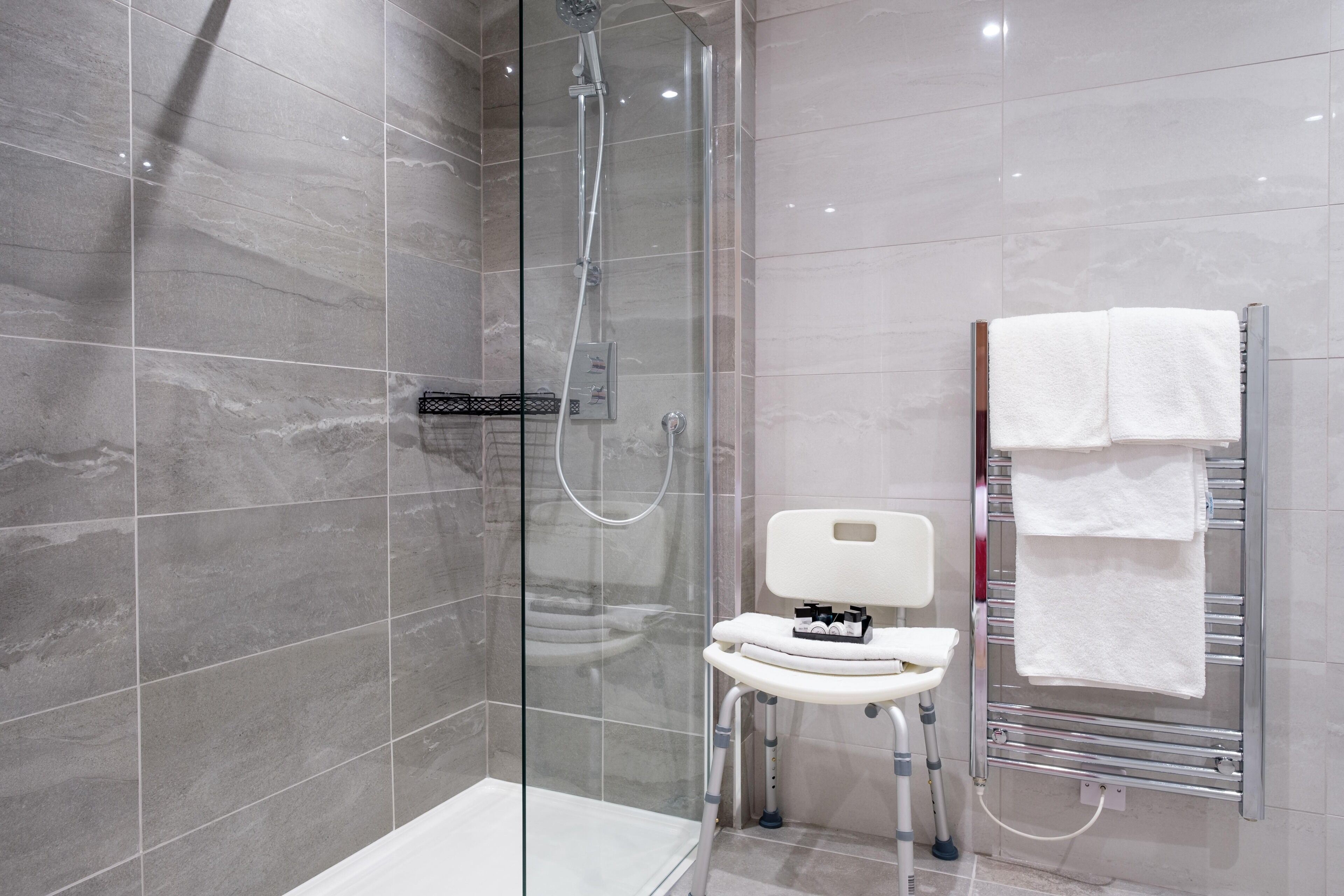 Un baño moderno con ducha envuelta en vidrio, inodoro blanco con mesita auxiliar y toallero con varias toallas.