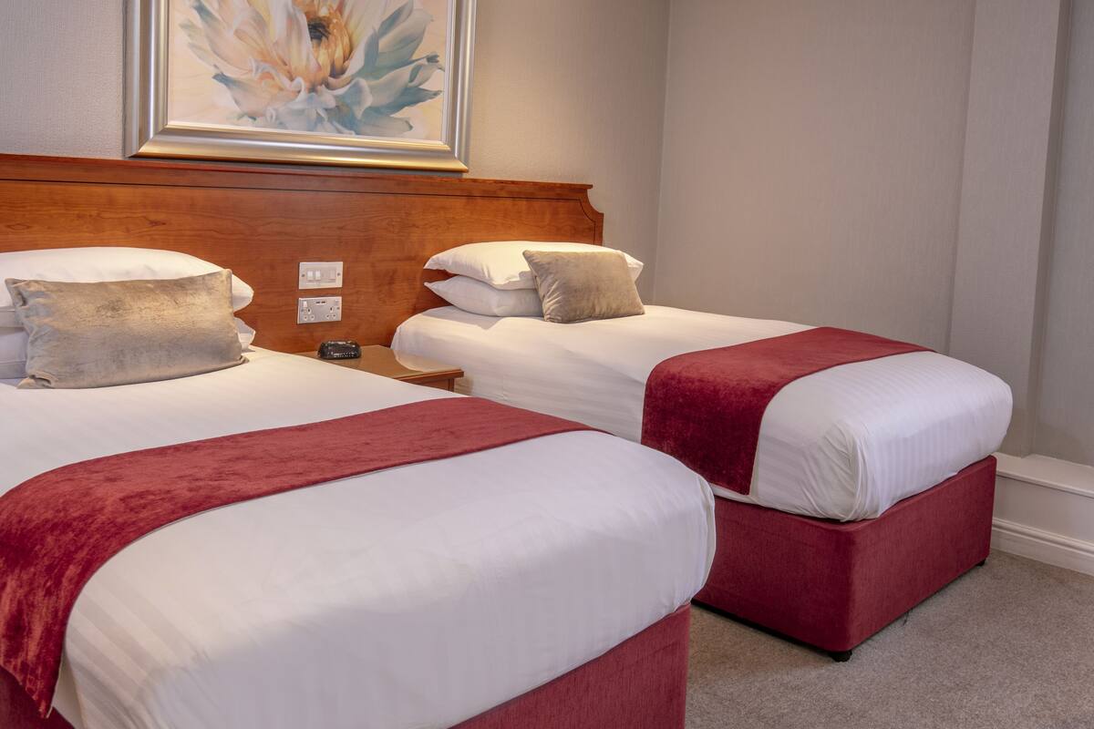 Habitación de hotel con dos camas, cada una con detalles en rojo, cabeceros de madera y un cuadro floral enmarcado sobre las camas.