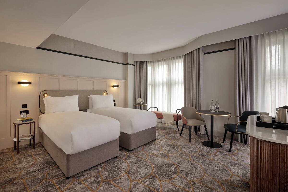 Une chambre d’hôtel avec deux lits, une petite table, une chaise, un bureau et une grande fenêtre avec des rideaux.