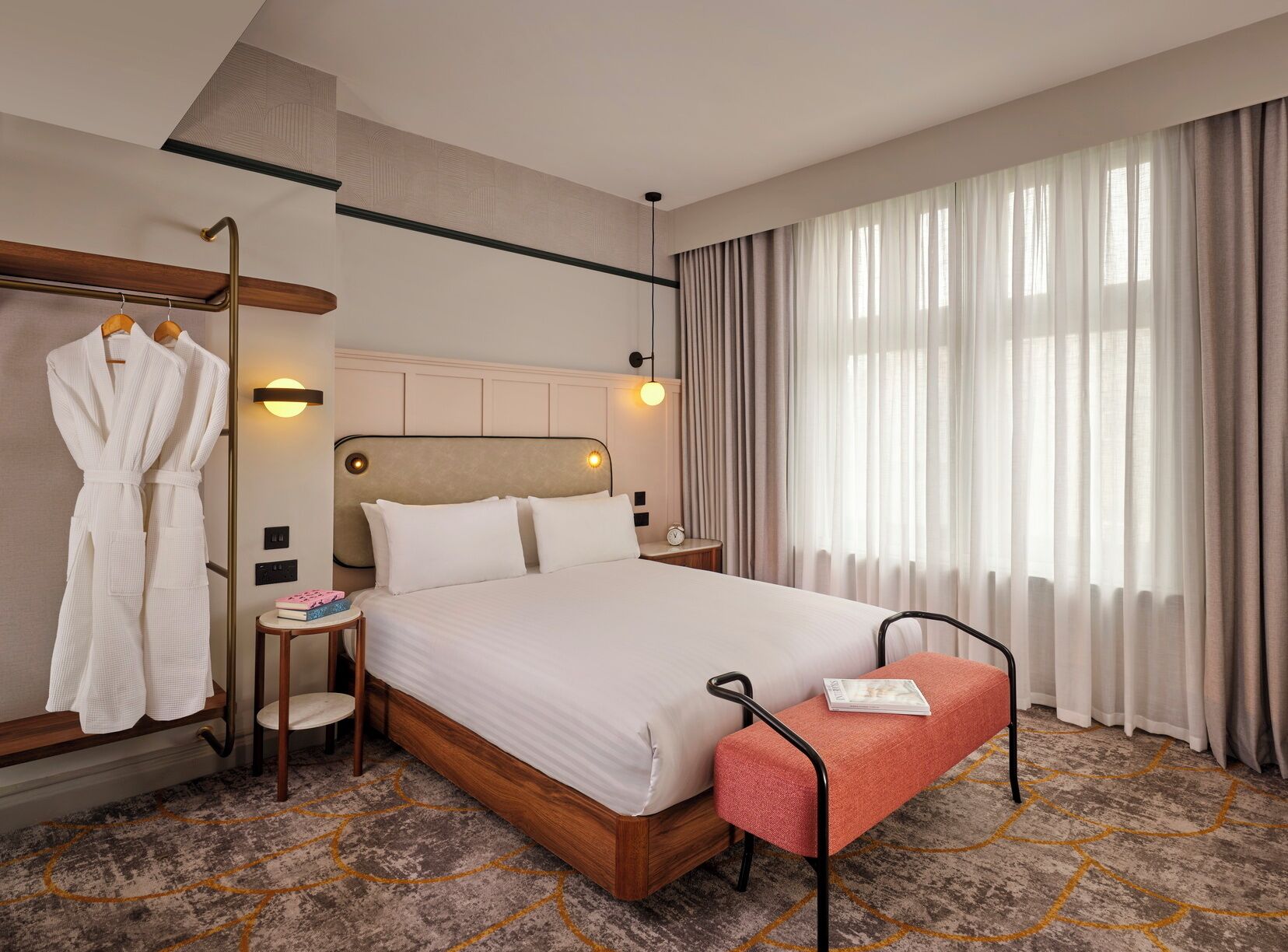 A hotel room with a bed, bedside tables, a robe rack, a bench, and a large window with curtains.