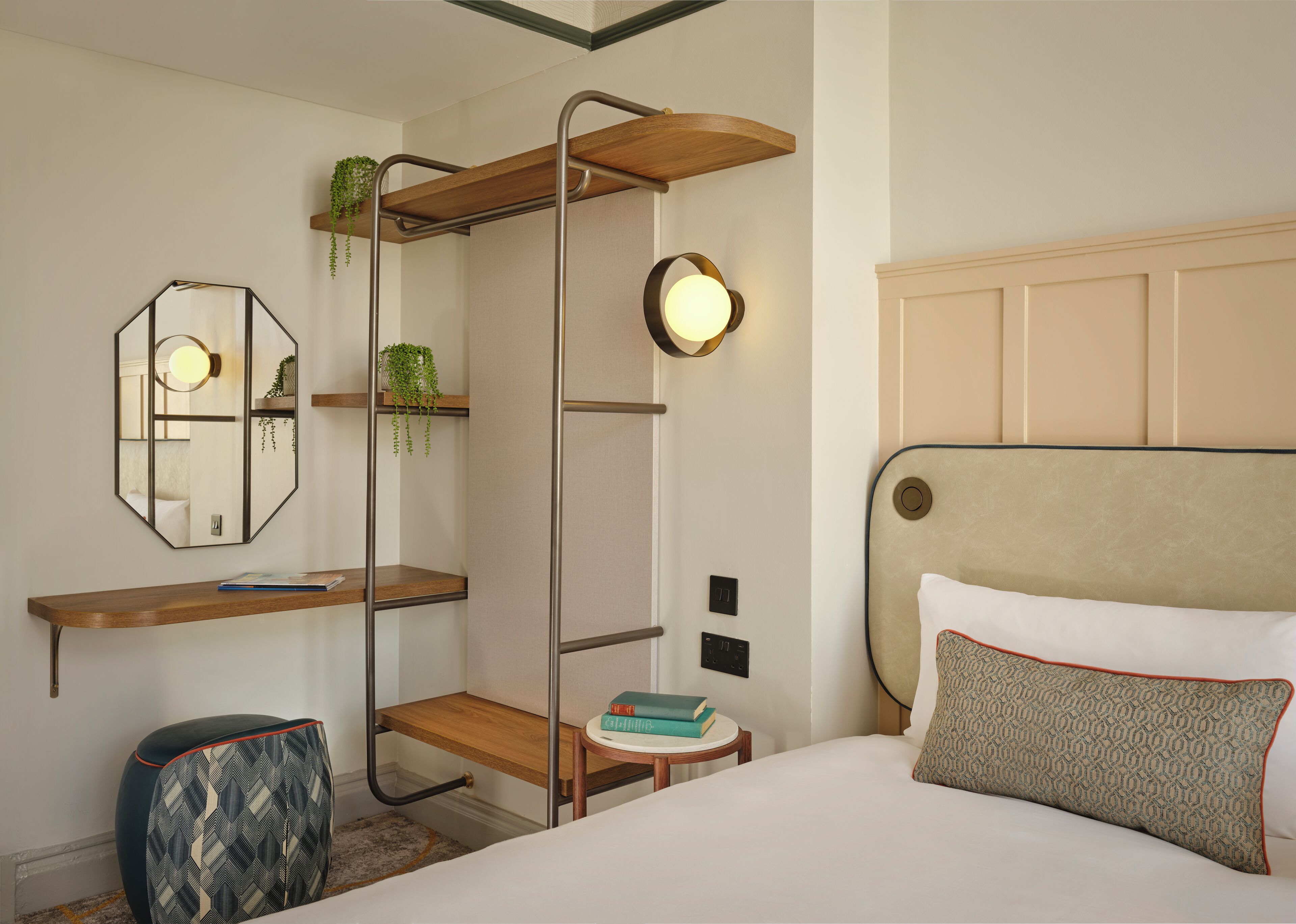 A hotel room with a bed, a bedside table with books, a mirror, a wall-mounted shelf with plants, and a hanging light fixture.
