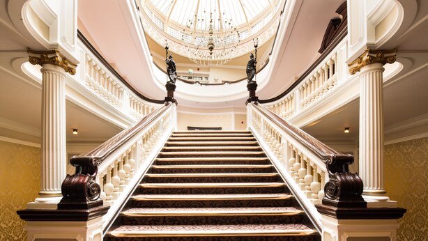 Un magnifique escalier, agrémenté d’un tapis à motifs, mène à un plafond haut orné d’un lustre.
