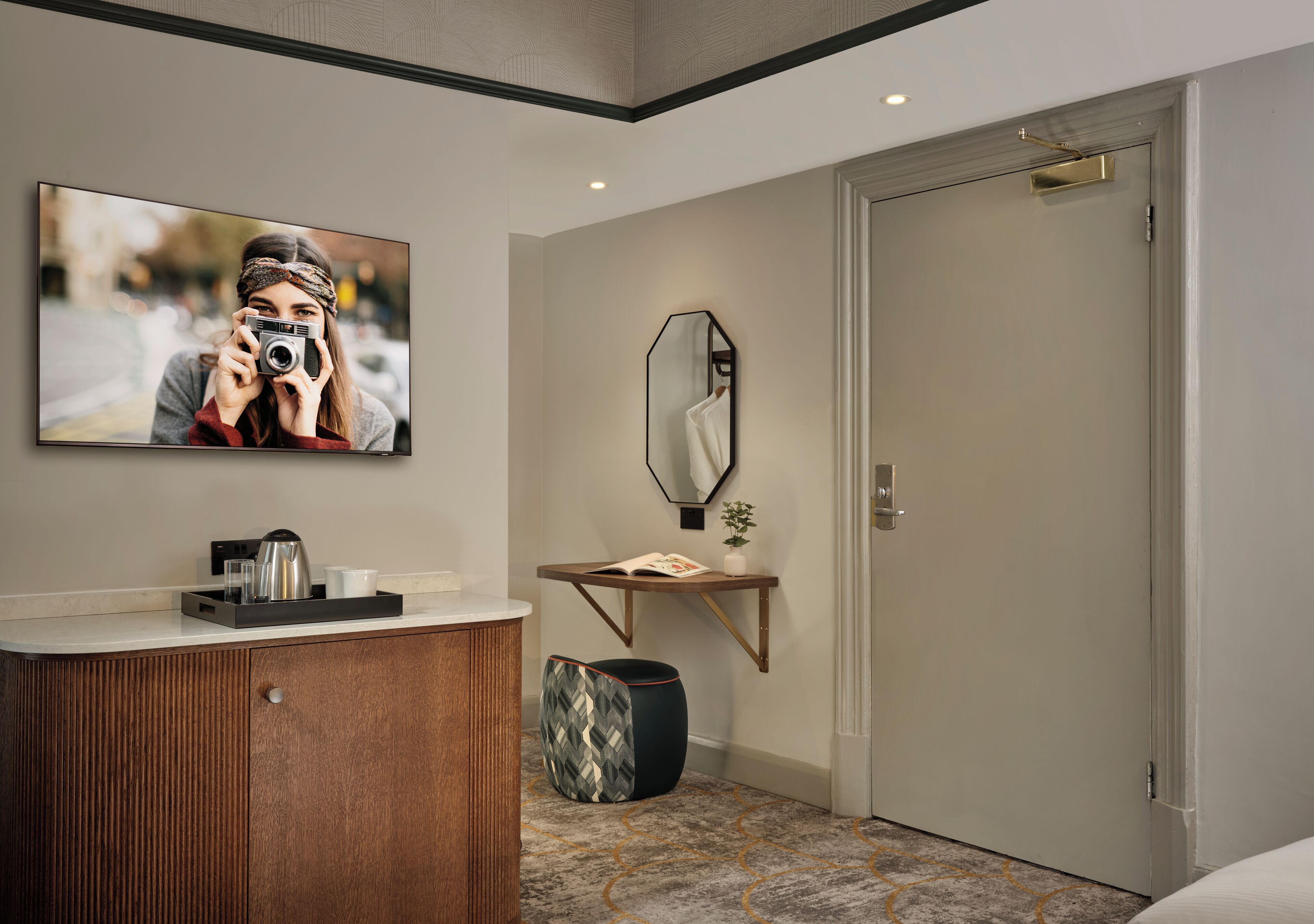 Ein modernes Hotelzimmer mit einem Holzkasten, einem Spiegel, einem kleinen Tisch mit Buch und Pflanze sowie einer Tür.
