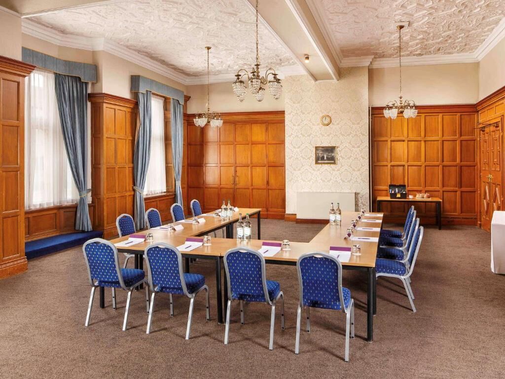 Un salón de conferencias con una larga mesa, sillas azules, paneles de madera, candelabros y suelo alfombrado.