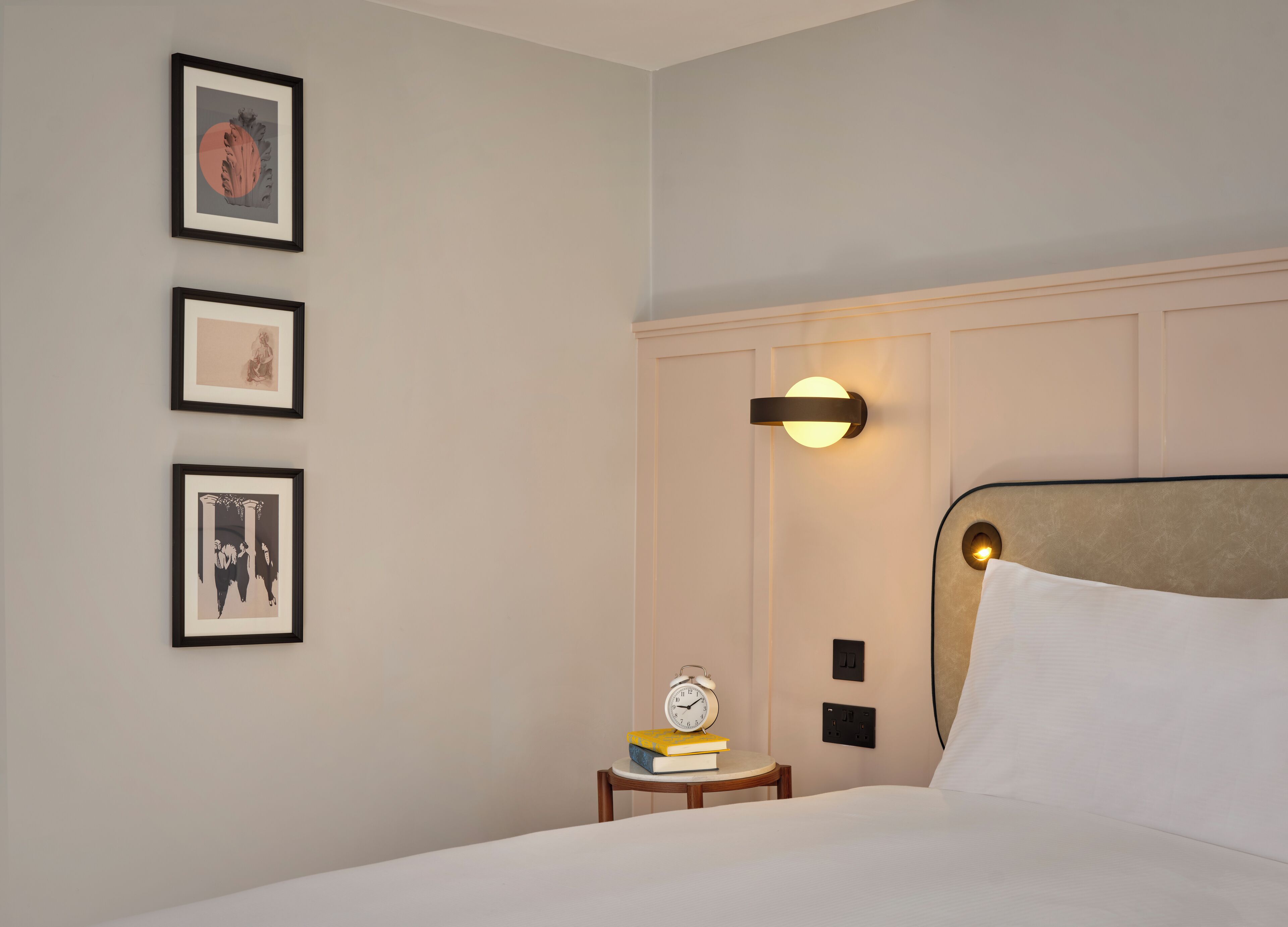 Een hotelkamer met een bed, nachtkastje met een klok en boeken, een wandlamp en een kunstwerk in een lijst aan de muur.