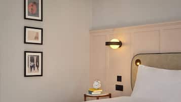 A hotel room with a bed, bedside table with a clock and books, a wall-mounted light fixture, and framed artwork on the wall.